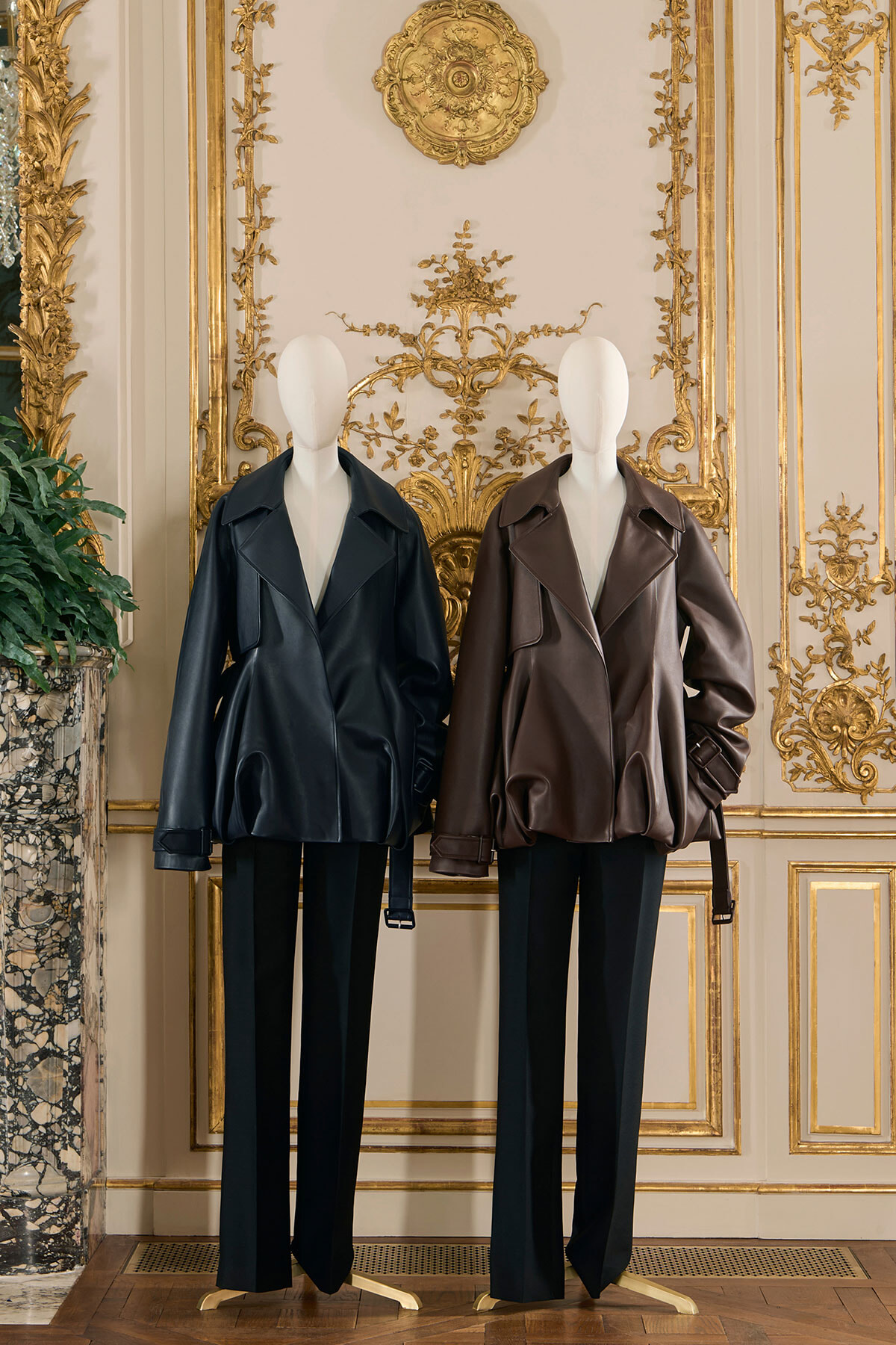 Two mannequins wearing oversized leather jackets (black and brown) on display in an ornate gilded room, black trousers, luxurious decor backdrop.