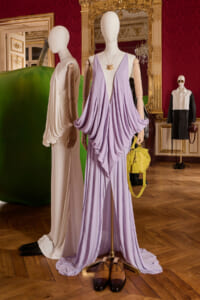 Mannequins display an elegant lavender draped gown with a yellow handbag in an ornate room showroom.