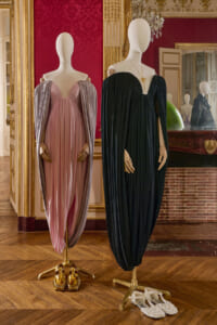 Two mannequins wear flowing evening gowns—pink-lavender on the left and black on the right—set in a gilded, ornate room with a red damask wall and mirror reflections.