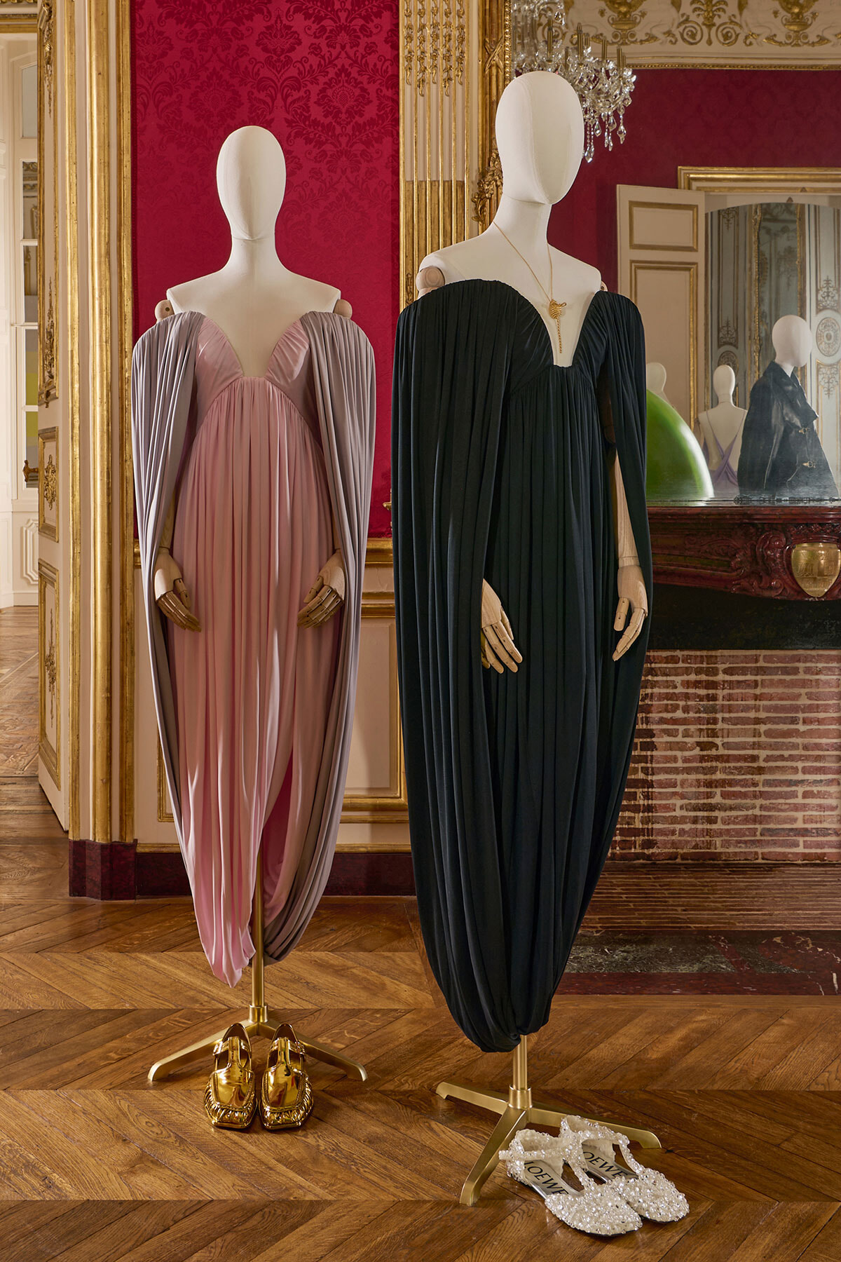 Two mannequins wear flowing evening gowns—pink-lavender on the left and black on the right—set in a gilded, ornate room with a red damask wall and mirror reflections.