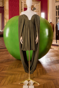 Mannequin wearing a dark green draped gown with cape sleeves, displayed beside a large green sphere in a gilded, ornate room with a wooden floor.