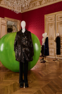 Mannequin wearing a black leather duffle coat in a luxurious room with red patterned walls and a large green sphere behind it.