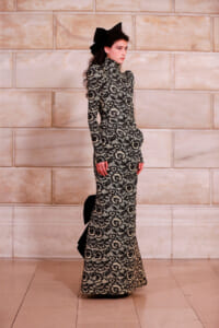 Model in a black-and-white lace gown with a high neck and long sleeves, standing in profile against a stone wall, wearing a large black bow in her hair.