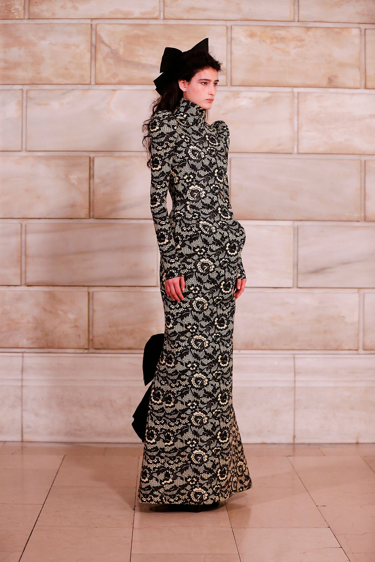 Model in a black-and-white lace gown with a high neck and long sleeves, standing in profile against a stone wall, wearing a large black bow in her hair.