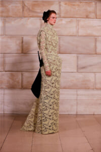 Model wearing a beige lace turtleneck gown with long sleeves and a black back bow, walking on a marble-tiled runway.