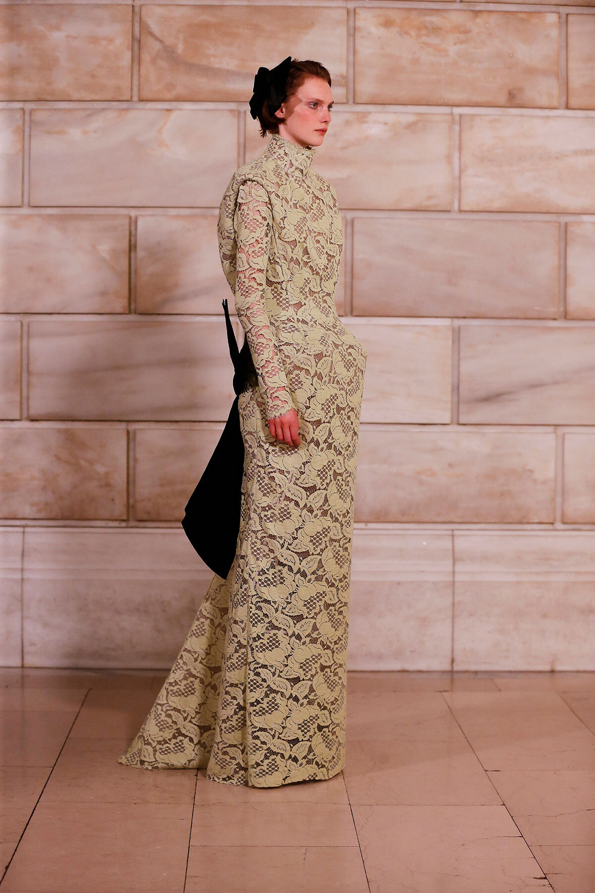 Model wearing a beige lace turtleneck gown with long sleeves and a black back bow, walking on a marble-tiled runway.