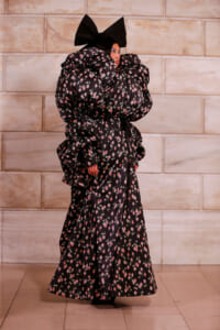 Model in a black floral-patterned gown with oversized puff sleeves and a large bow headpiece, standing against a stone wall.