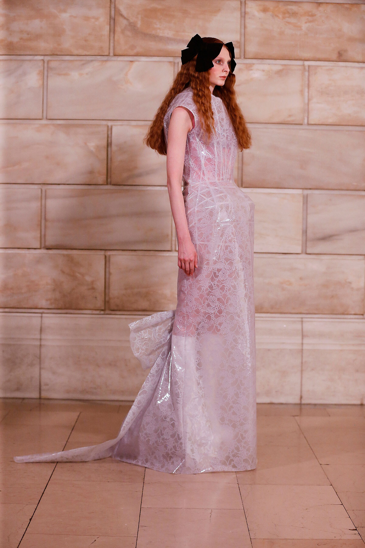 Model in a lavender lace evening gown with a long train, standing sideways in a marble-walled room, hair adorned with large black bows.