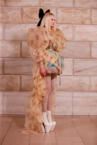 Model wearing a sheer, floral gown with oversized puff sleeves, tied at the waist, paired with white platform boots and a large black bow in her hair.