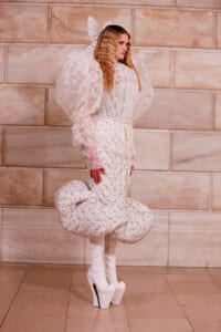 Model in a white floral lace dress with dramatic puff sleeves and white platform boots, posing against a beige stone wall.