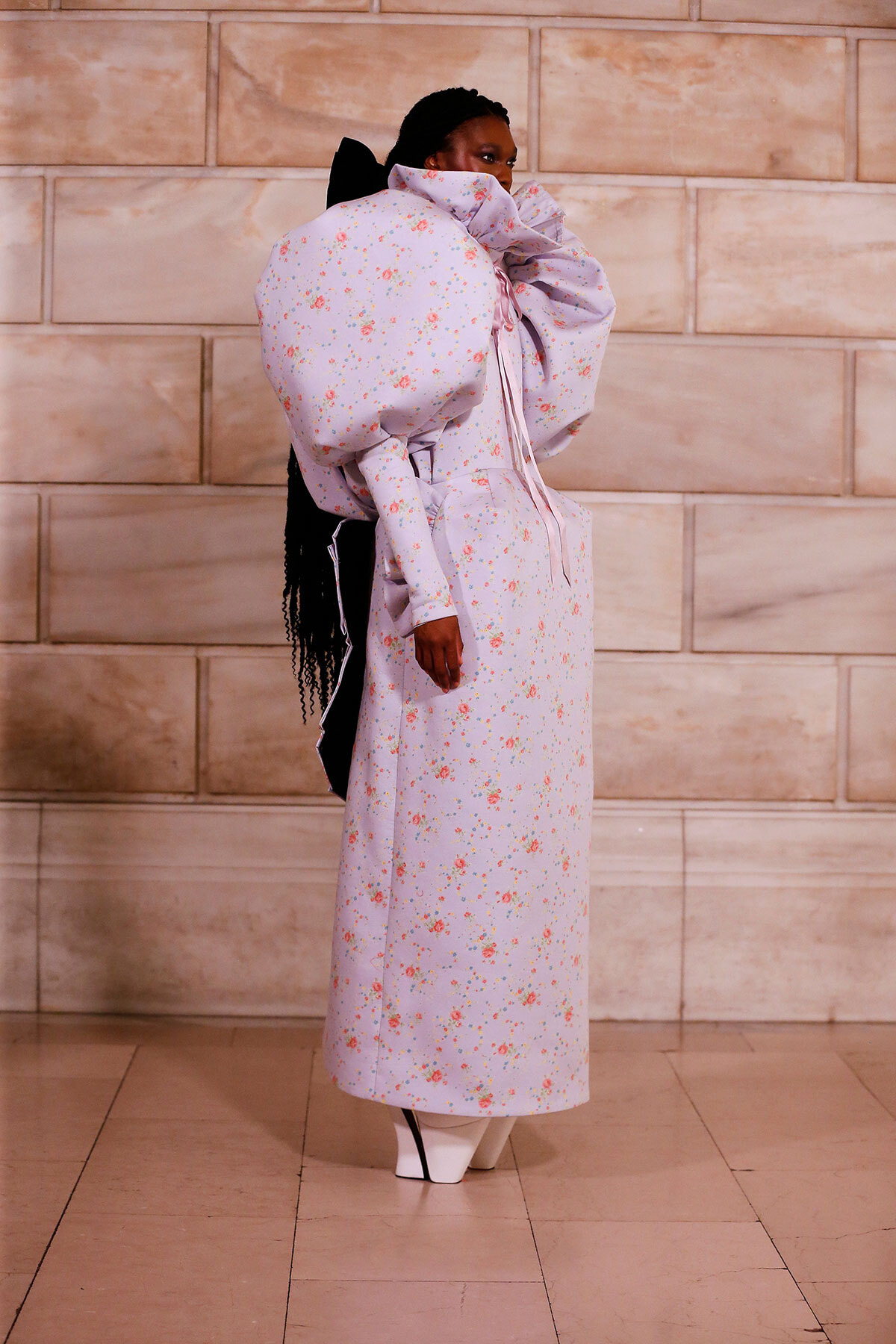 Model in a lavender floral gown with oversized puff sleeves and a long skirt, posing against a beige stone wall.