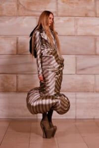 Model wearing a metallic striped avant-garde dress with oversized hip poufs and platform heels, posed against a stone wall backdrop.