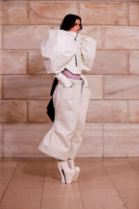 Model in a sculptural white floral blouse with oversized sleeves, tucked into high-waisted beige wide-leg pants and white platform heels, indoors against a stone wall.