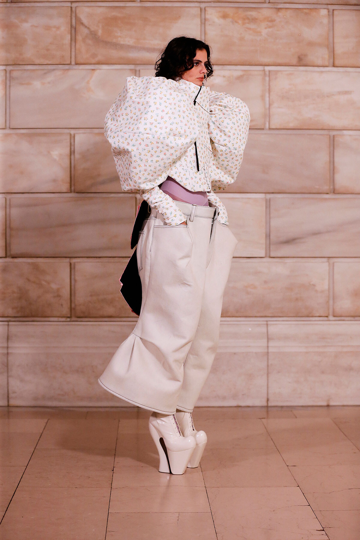 Model in a sculptural white floral blouse with oversized sleeves, tucked into high-waisted beige wide-leg pants and white platform heels, indoors against a stone wall.