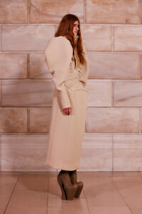 Side view of a model in a beige oversized-sleeve coat and platform boots, standing against a stone wall.