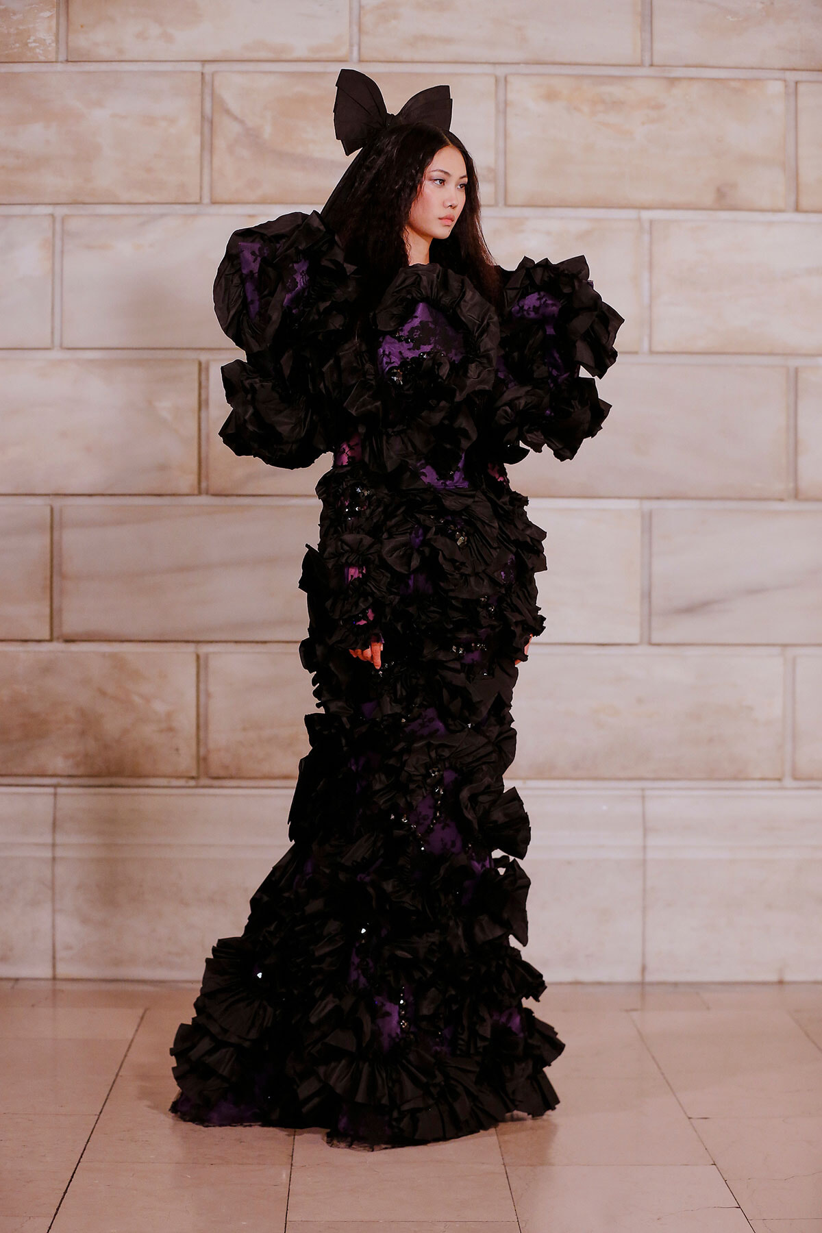 Model wearing a dramatic black ruffled gown with purple accents and a large bow headpiece, standing against a stone wall indoors.