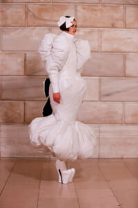 Model walks the runway in an all-white sculptural gown with oversized puff sleeves and a bulbous, tiered lower skirt, paired with white platform heels.