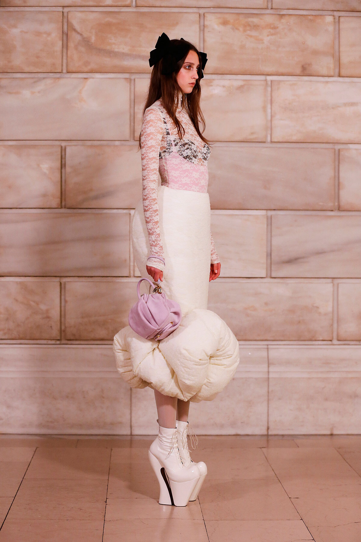 Side-profile fashion model wearing a lace pink top and white skirt, with black bows, carrying a lavender handbag and a large cream sculptural accessory, in white platform boots against a stone wall.