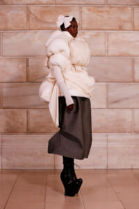 Model wearing oversized white puffer top with sculptural sleeves and a large bow headband, paired with a gray midi skirt and black platform boots against a stone wall backdrop.