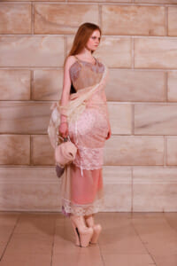 Model in pale pink lace dress with beige overlay, carrying a small pink bag, posing against a stone wall backdrop.