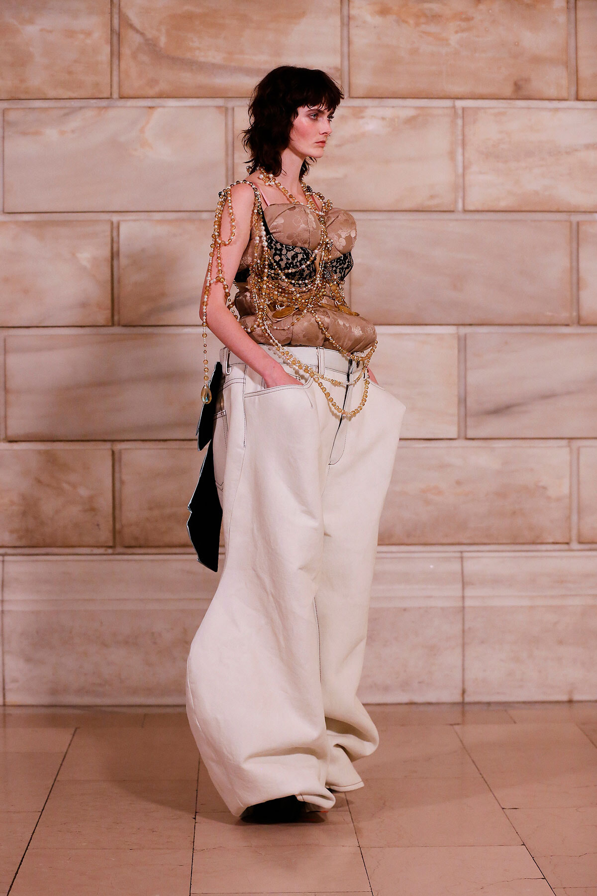 Runway model wearing a nude-toned lacy corset top with gold bead chains and oversized white trousers against a stone wall background.