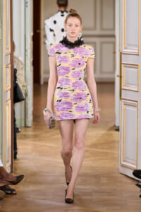 Model walking a runway in a short pastel pink and yellow ruched dress with a black lace collar, black heels, and a small metallic clutch.