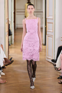 Model walking a runway in a pastel pink lace sheath dress, black lace tights, and white polka-dotted heels in a gilded room.