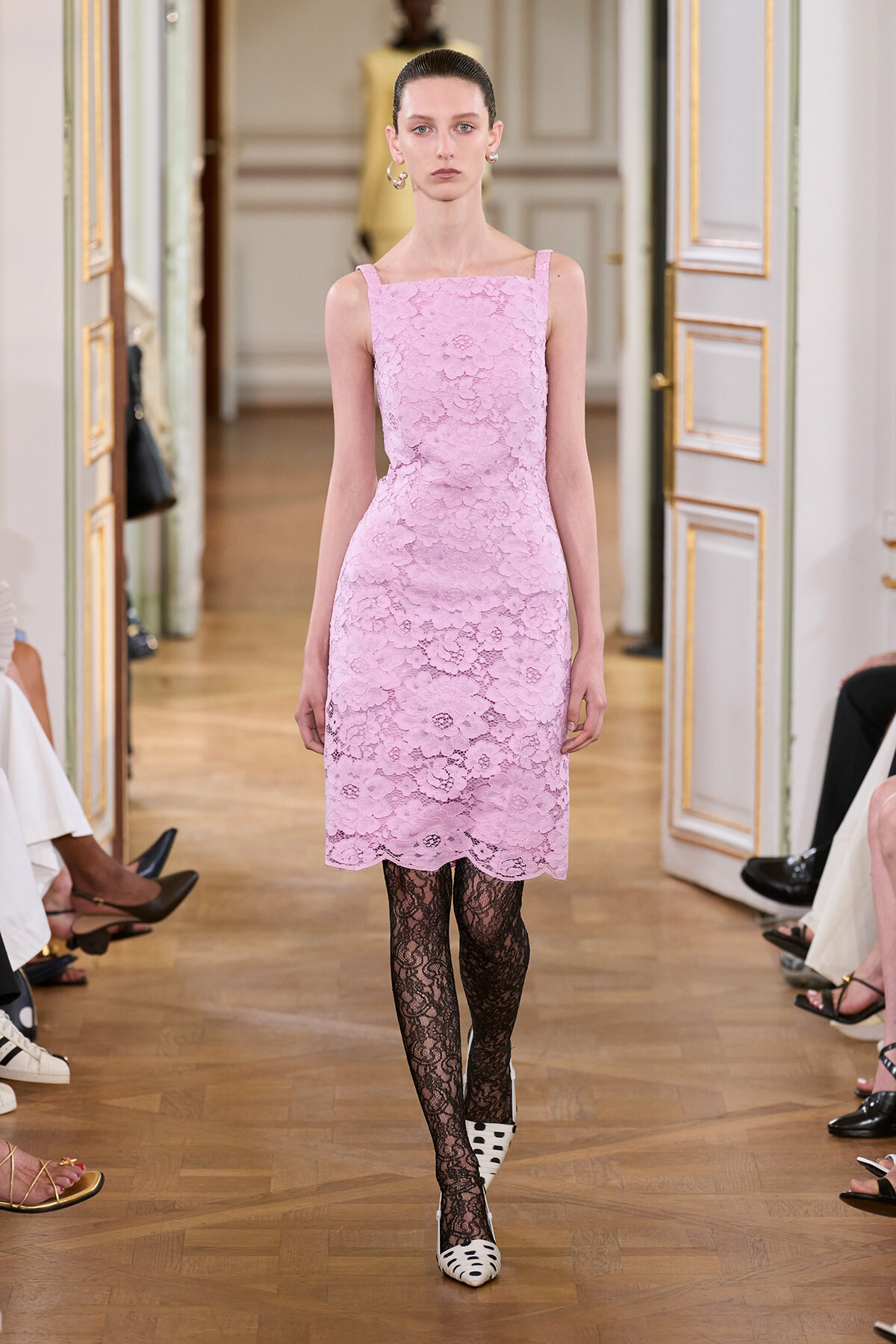 Model walking a runway in a pastel pink lace sheath dress, black lace tights, and white polka-dotted heels in a gilded room.