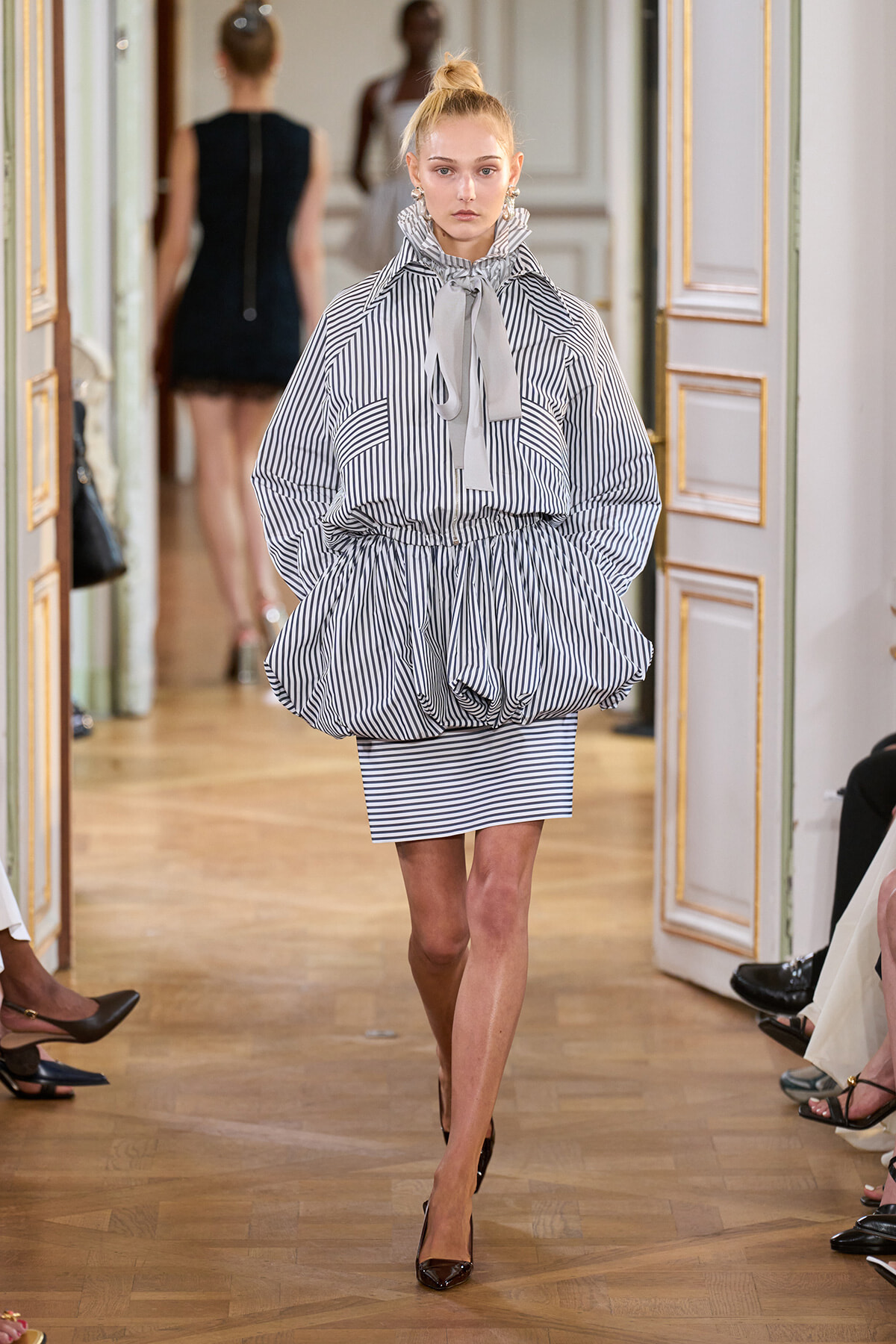 Model walks a fashion runway in a navy-and-white striped outfit with a voluminous bubble skirt and bow-tied neck scarf.