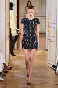 Model walking the runway in a black polka-dot mini dress with ruching and a pearl choker, wearing a cat-ear headband in a gilded hallway setting.
