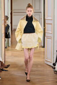 Model walking the runway in a pale yellow trench over a black lace crop top and cream lace mini skirt at a fashion show.