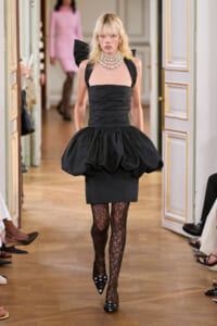 Model walking a runway in a black strapless dress with a voluminous bubble skirt and stacked pearl necklace, lime-green walls in a gilded room setting.