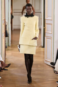 Model walking a runway in a pale yellow tailored coat and matching skirt, with a black lace collar and black tights.