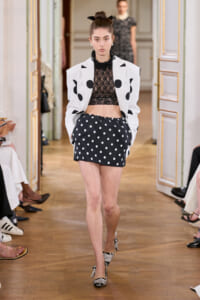 Model walking a fashion show in a white blazer with black polka dots, a black lace crop top, and a navy polka-dot mini skirt; hair in a bun with a bow.