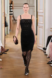 Model walking a runway in a black lace midi dress with thin straps, carrying a small round burgundy handbag.