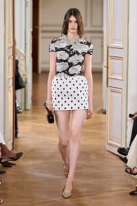 Model walks a runway in a black-and-white abstract-print top and white polka-dot mini skirt, holding a small black clutch.