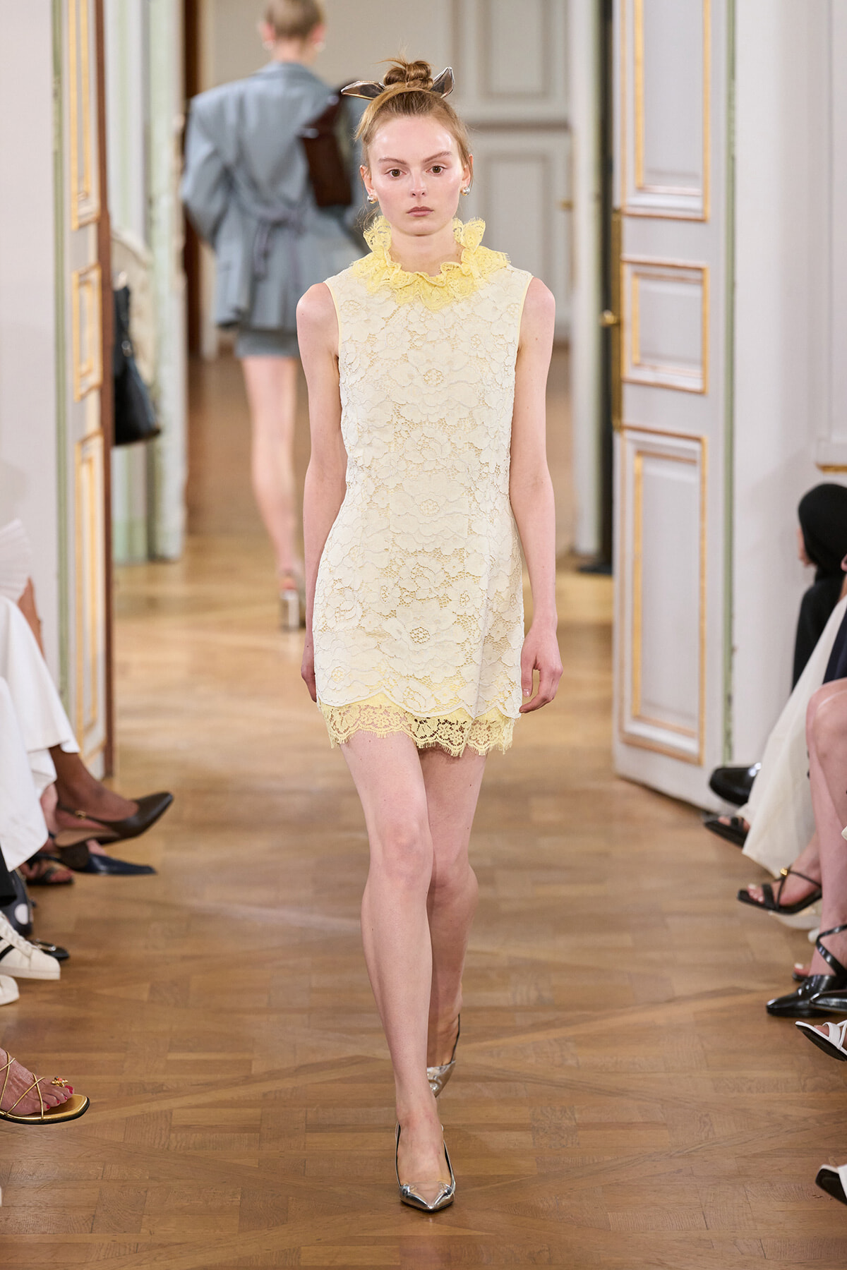 Female model walking a runway in a pale yellow lace sleeveless dress with a ruffled high neck collar.
