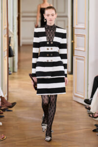 Model walks a runway wearing a black-and-white striped double-breasted blazer dress with a lace high neck and patterned tights.
