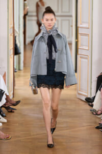 Model walks the runway wearing a light blue oversized jacket over a striped ruffled blouse and black lace mini skirt, with black patent heels.