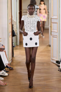 Runway model walks the catwalk in a white polka-dot top and mini skirt with large black spots, plus a chunky pearl necklace.