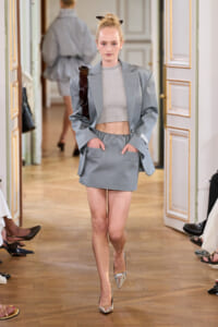 Model walks a runway in a light gray oversized blazer with matching mini skirt and cropped top, silver metallic heels.