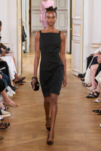 Model walking a runway in a black sleeveless dress, holding a small round black clutch, hair tied with a scarf.