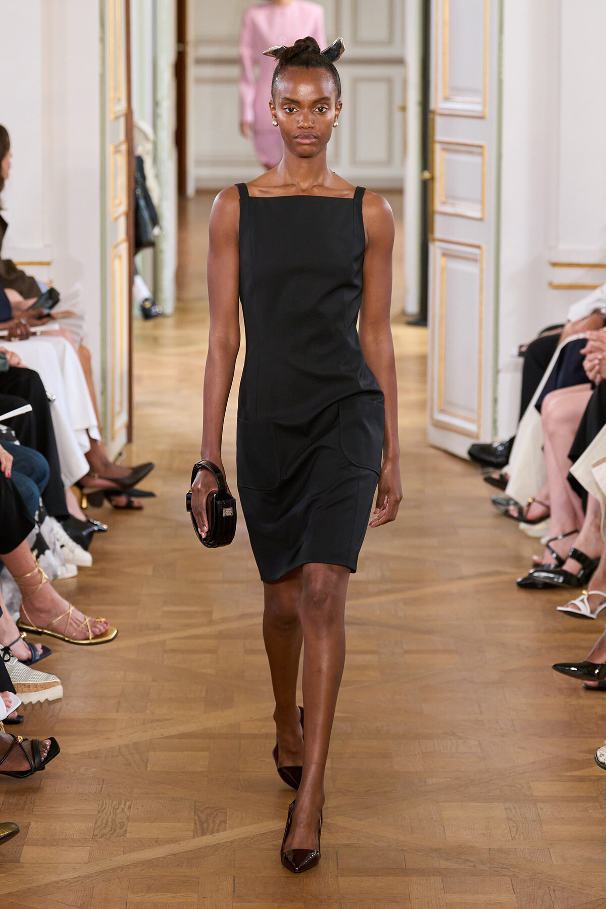 Model walking a runway in a black sleeveless dress, holding a small round black clutch, hair tied with a scarf.