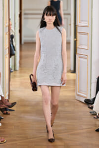 Model walking a runway in a light gray lace sleeveless mini dress, holding a dark brown clutch and wearing black heels.