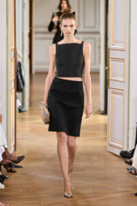 Model walking a runway in a black ruched crop top and knee-length black skirt, holding a metallic clutch.
