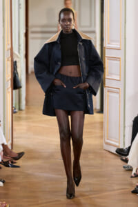 Model walks a runway in a navy oversized coat with a wide beige collar, paired with a black crop top and navy mini skirt, hands in coat pockets.