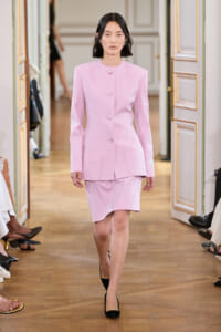 Model walking a runway in a pale pink tailored blazer with matching skirt and black heels in an ornate venue.