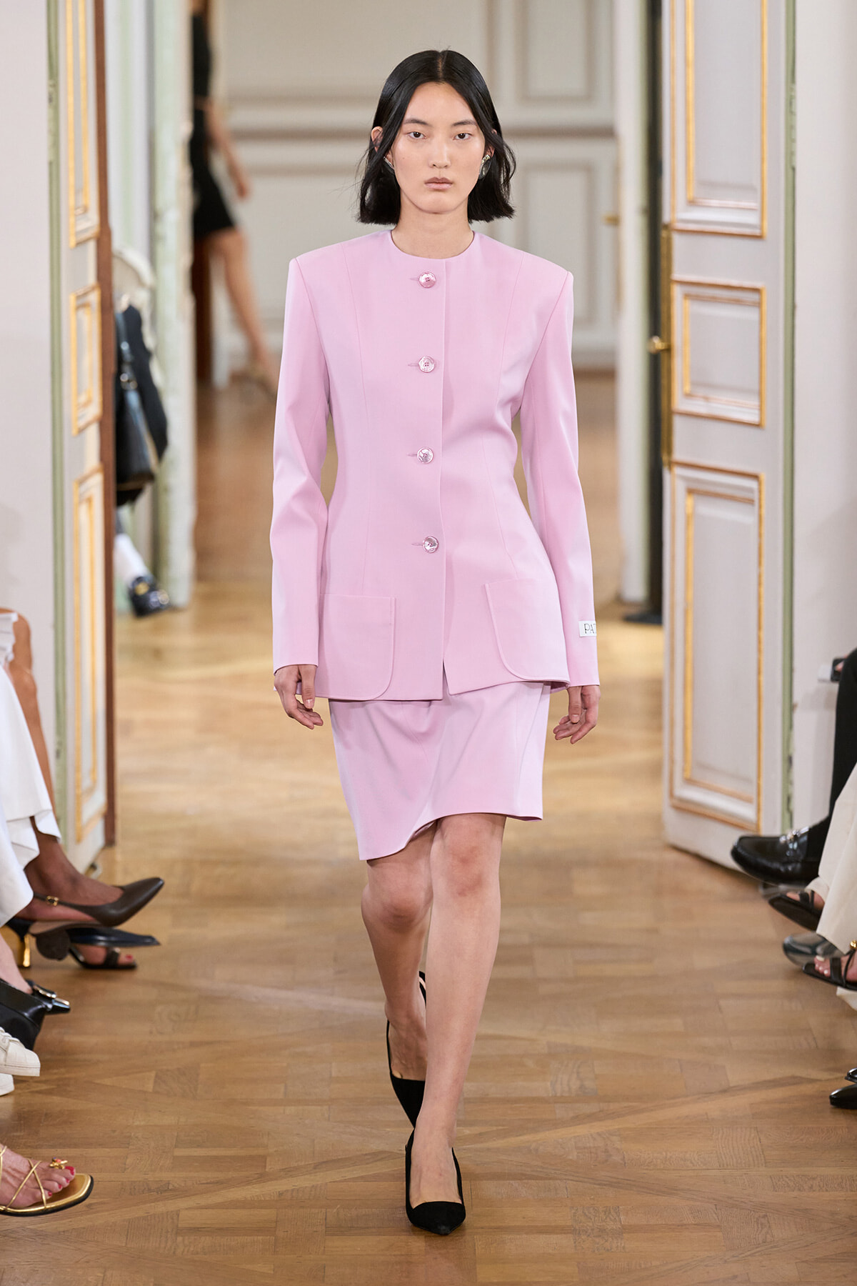 Model walking a runway in a pale pink tailored blazer with matching skirt and black heels in an ornate venue.