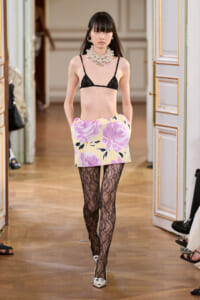Runway model wearing a black lace bralette, yellow floral mini skirt, black lace tights, white shoes, and a chunky pearl necklace.