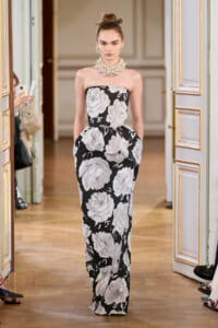 Model wearing a strapless black-and-white floral gown with a voluminous skirt and a chunky pearl necklace, walking in an ornate doorway hallway.